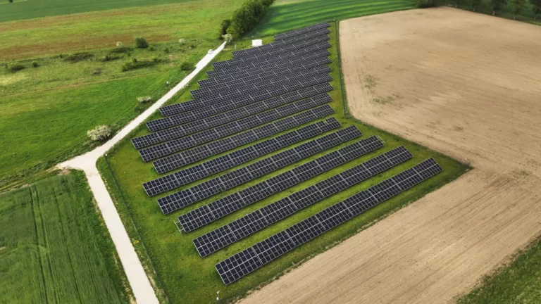 Farma fotowoltaiczna na dużym areale rolnym z widoczną w oddali turbiną wiatrową – przykład inwestycji w odnawialne źródła energii.