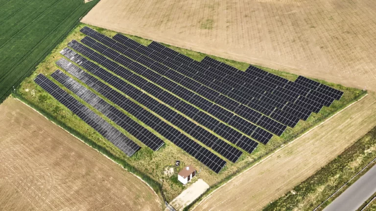 Instalacja paneli fotowoltaicznych ułożona na lekko wzniesionym terenie rolnym, otoczona zielonym krajobrazem.